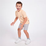 A young boy wearing a light peach polo shirt, light gray shorts, gray socks, and white sneakers, posing in a playful crouched stance against a solid white studio background.