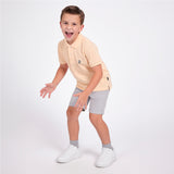 A young boy wearing a light peach polo shirt, light gray shorts, gray socks, and white sneakers, posing in a playful crouched stance against a solid white studio background.