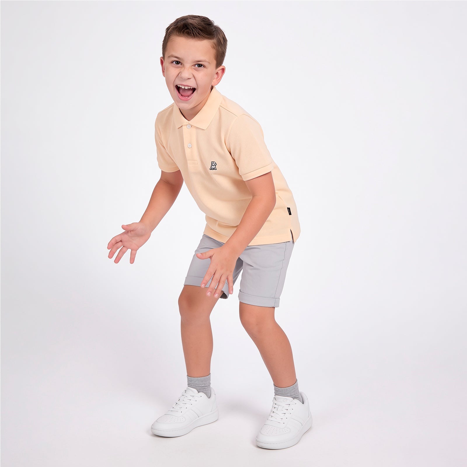 A young boy wearing a light peach polo shirt, light gray shorts, gray socks, and white sneakers, posing in a playful crouched stance against a solid white studio background.