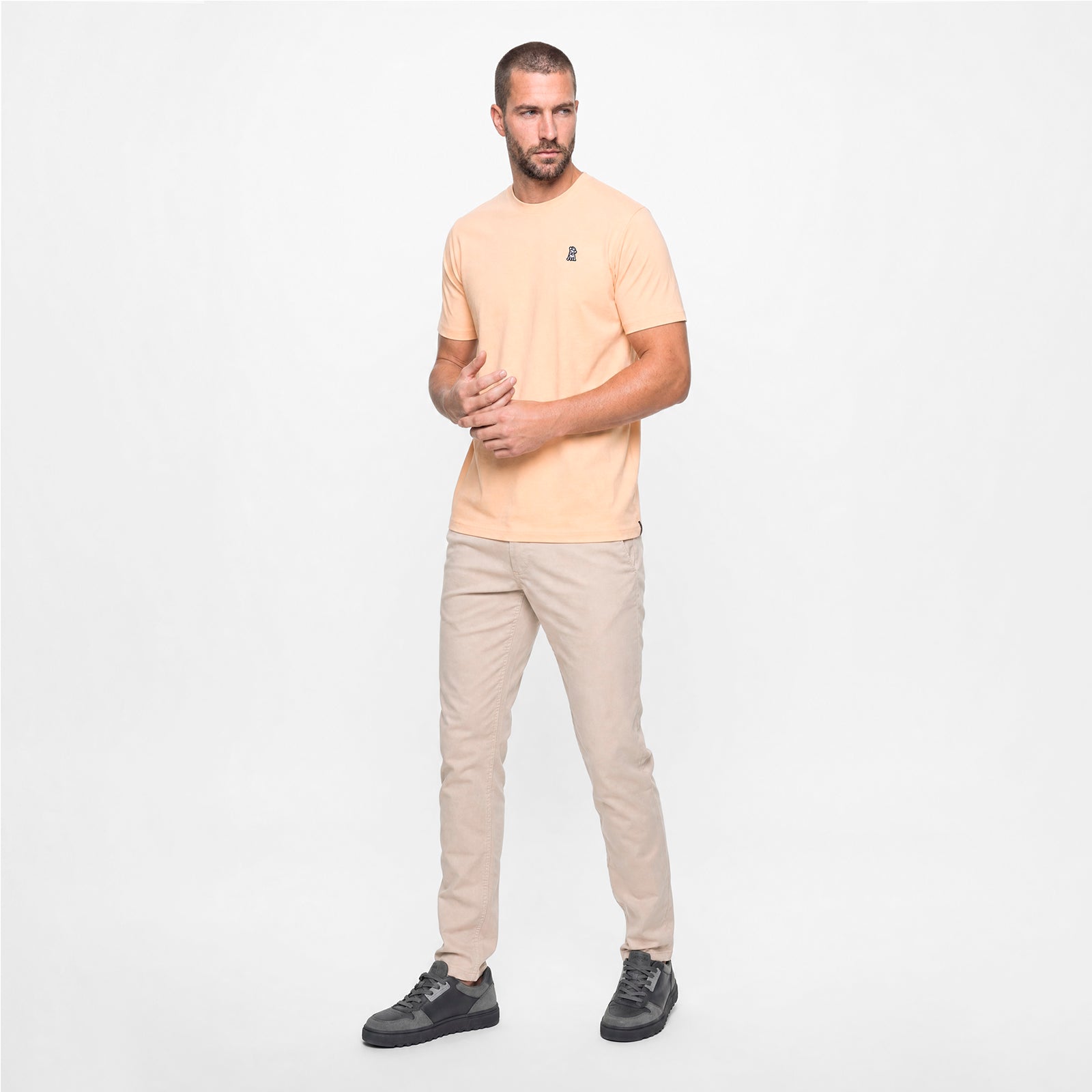 A man wearing a peach color t-shirt with beige pants on a white background