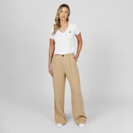 Full-length front view of a female model wearing a white v-neck t-shirt with a gold logo, wide-leg beige trousers, and white sneakers, standing against a light grey background