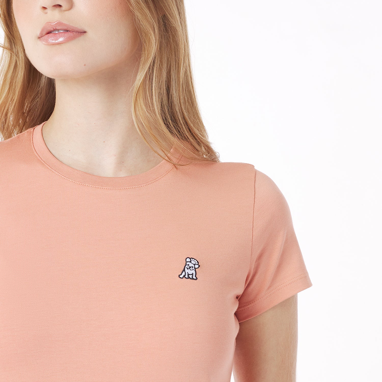 Close-up of a woman wearing a peach-colored t-shirt with a small logo on a white background