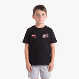 A young boy wearing a black short-sleeve t-shirt with two small racing-style chest patches and light gray shorts, standing against a plain light gray background.