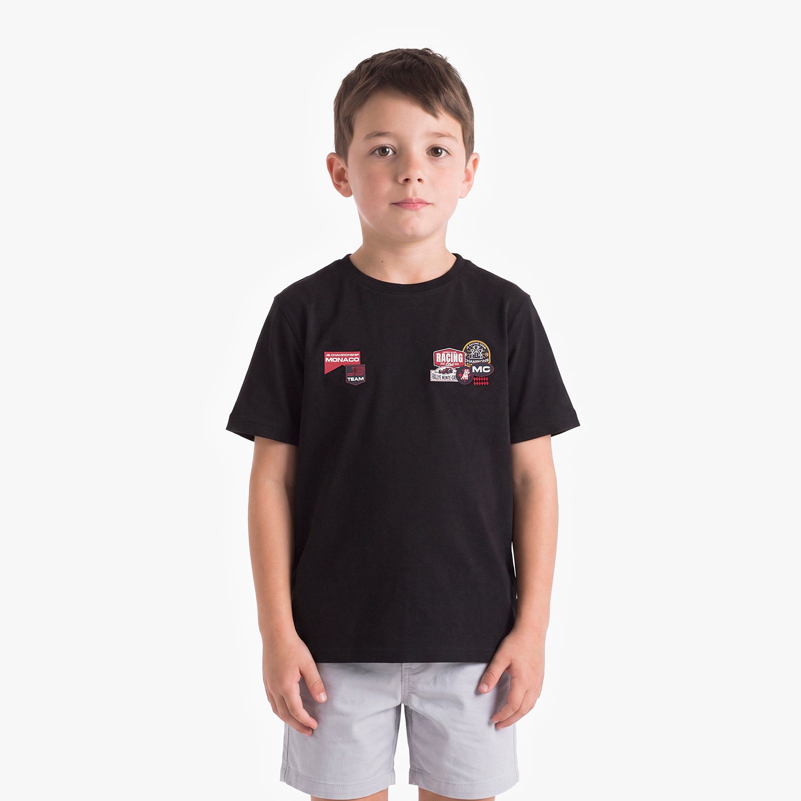 A young boy wearing a black short-sleeve t-shirt with two small racing-style chest patches and light gray shorts, standing against a plain light gray background.