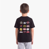 A child viewed from behind wearing a black short-sleeve T-shirt with a collage of racing-themed patch graphics on the back, paired with light gray pants, against a plain light gray studio background.
