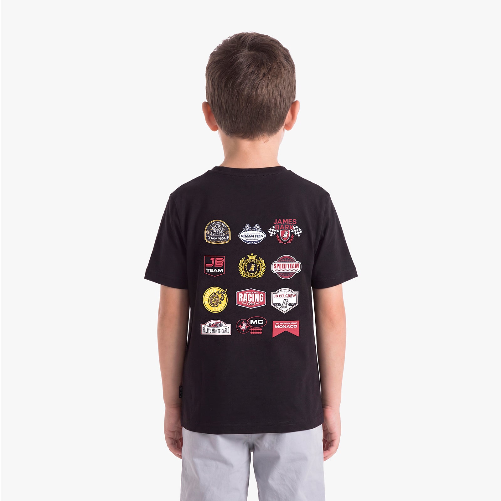 A child viewed from behind wearing a black short-sleeve T-shirt with a collage of racing-themed patch graphics on the back, paired with light gray pants, against a plain light gray studio background.