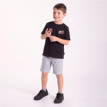 A young boy modeling a black short-sleeve t-shirt with a graphic patch on the left chest, paired with light gray shorts and black sneakers, standing against a plain white studio background.