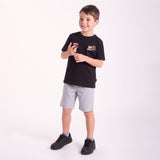 A young boy modeling a black short-sleeve t-shirt with a graphic patch on the left chest, paired with light gray shorts and black sneakers, standing against a plain white studio background.