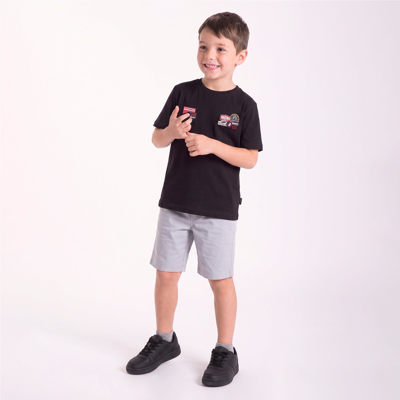A young boy modeling a black short-sleeve t-shirt with a graphic patch on the left chest, paired with light gray shorts and black sneakers, standing against a plain white studio background.