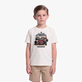 A young boy modeling a cream-colored t-shirt with a James Barker racing graphic, paired with khaki shorts