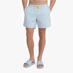 A model wearing light blue swim shorts with a yellow drawstring and a small yellow logo on the left leg, paired with matching light blue slide sandals