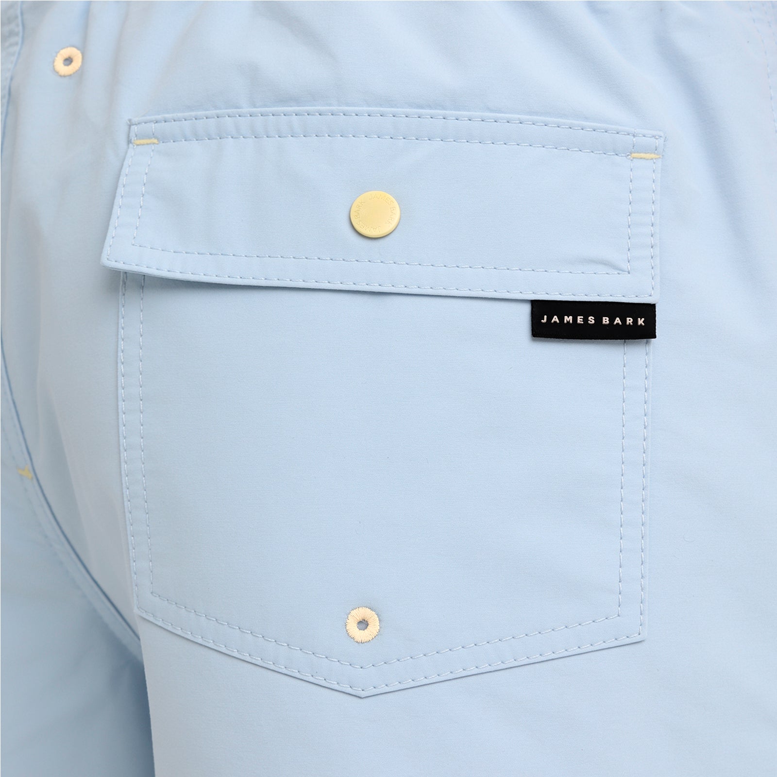 Close-up of a light blue shirt pocket with gold-tone snap buttons and a black James Bark brand label.