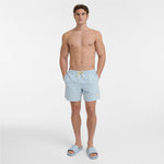 A shirtless male model wearing light blue swim shorts with a yellow drawstring and matching light blue slide sandals