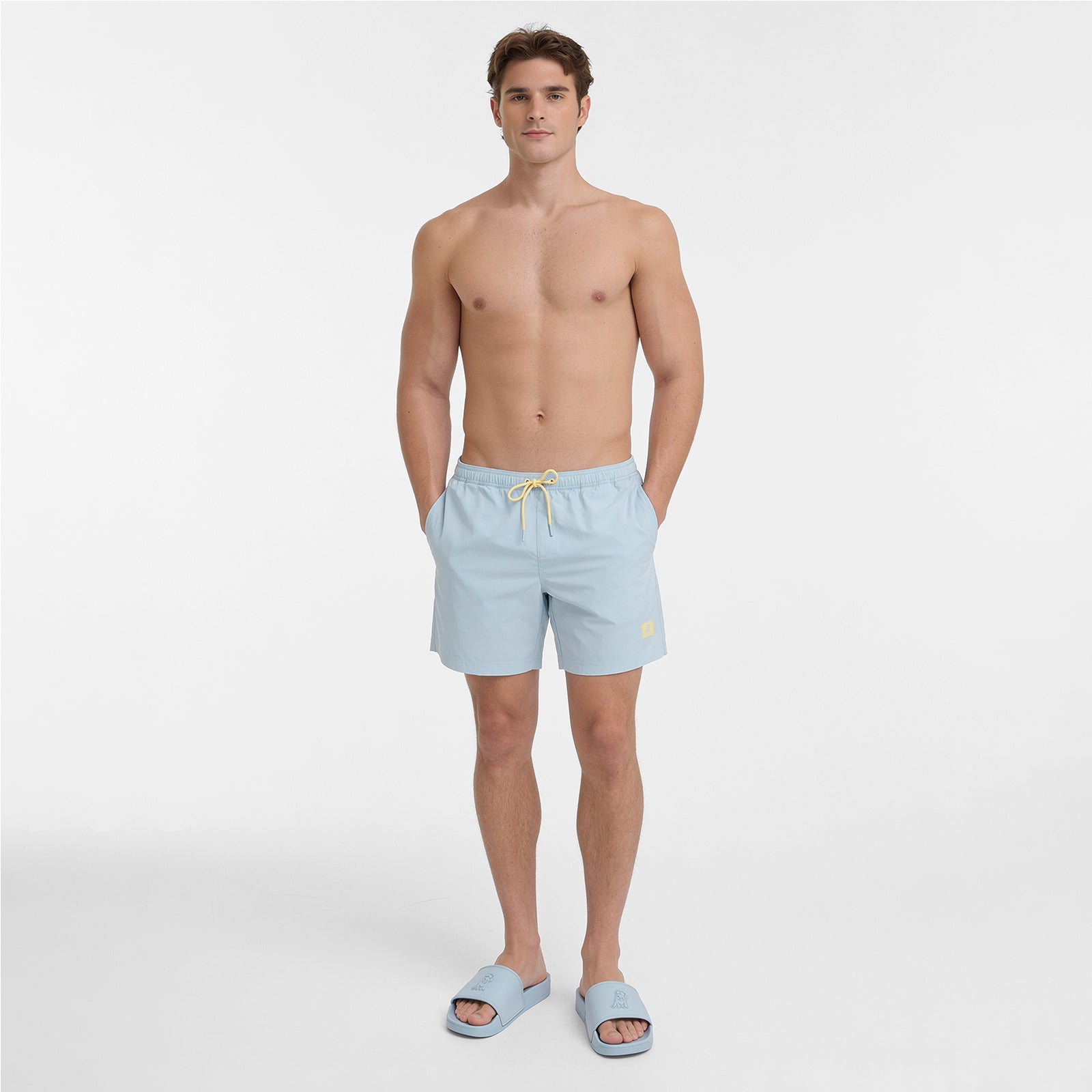 A shirtless male model wearing light blue swim shorts with a yellow drawstring and matching light blue slide sandals