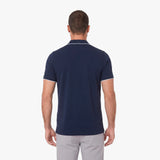 A man viewed from the back wearing a navy blue short-sleeve polo shirt with thin white piping trim on the collar and sleeve cuffs, paired with light gray trousers, against a plain white background.