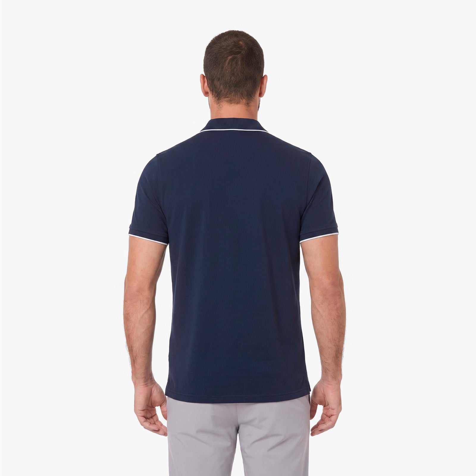 A man viewed from the back wearing a navy blue short-sleeve polo shirt with thin white piping trim on the collar and sleeve cuffs, paired with light gray trousers, against a plain white background.