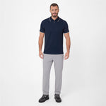 A man modeling a short-sleeve polo shirt in navy blue with white trim on the collar and sleeve cuffs, paired with light gray trousers and black sneakers, against a plain light gray studio background.