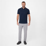 A man modeling a short-sleeve polo shirt in navy blue with white trim on the collar and sleeve cuffs, paired with light gray trousers and black sneakers, against a plain light gray studio background.