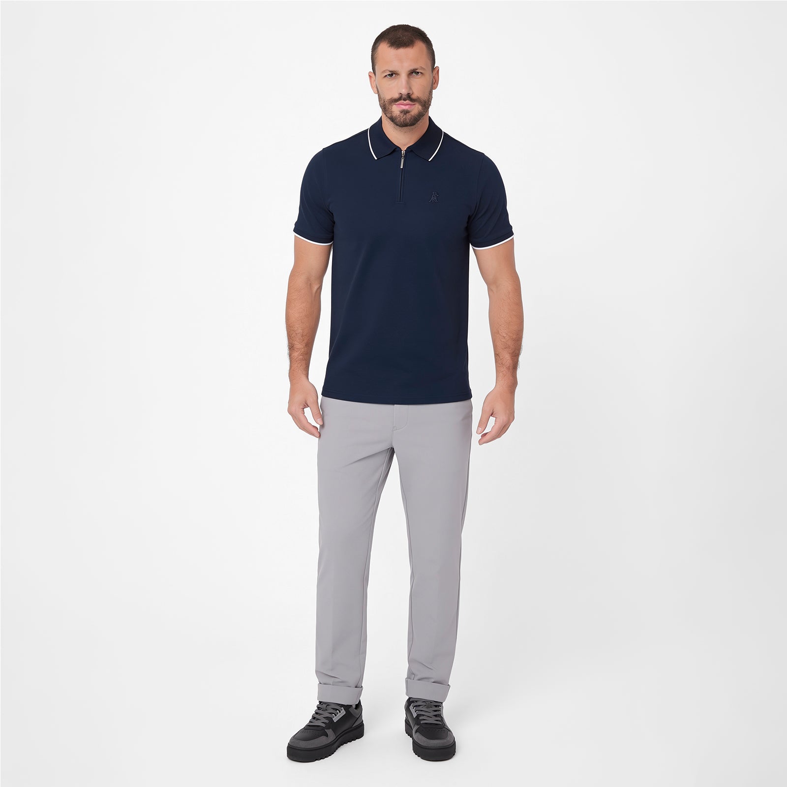 A man modeling a short-sleeve polo shirt in navy blue with white trim on the collar and sleeve cuffs, paired with light gray trousers and black sneakers, against a plain light gray studio background.