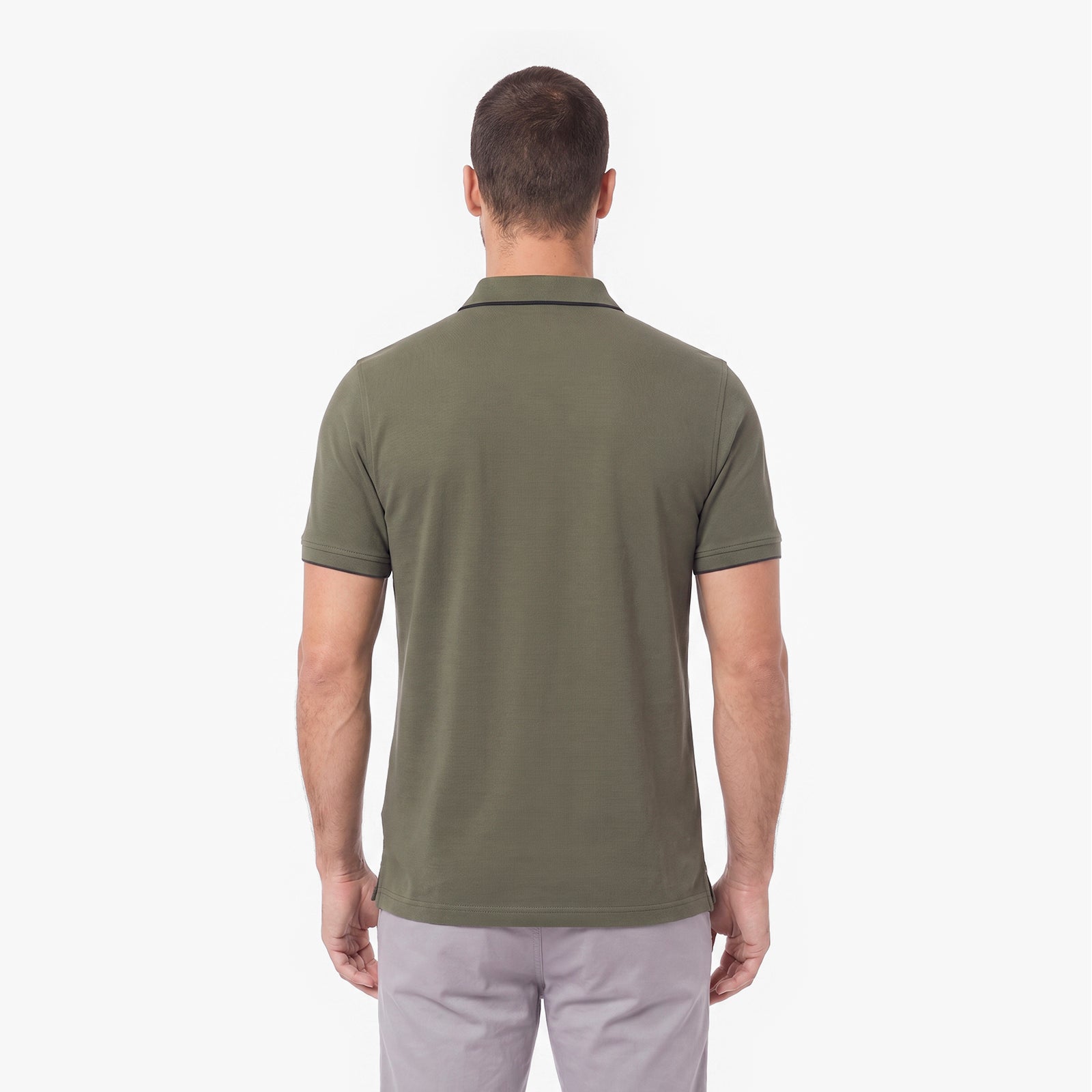 A man viewed from the back wearing an olive green short-sleeve polo shirt and light gray trousers, standing against a plain white studio background.