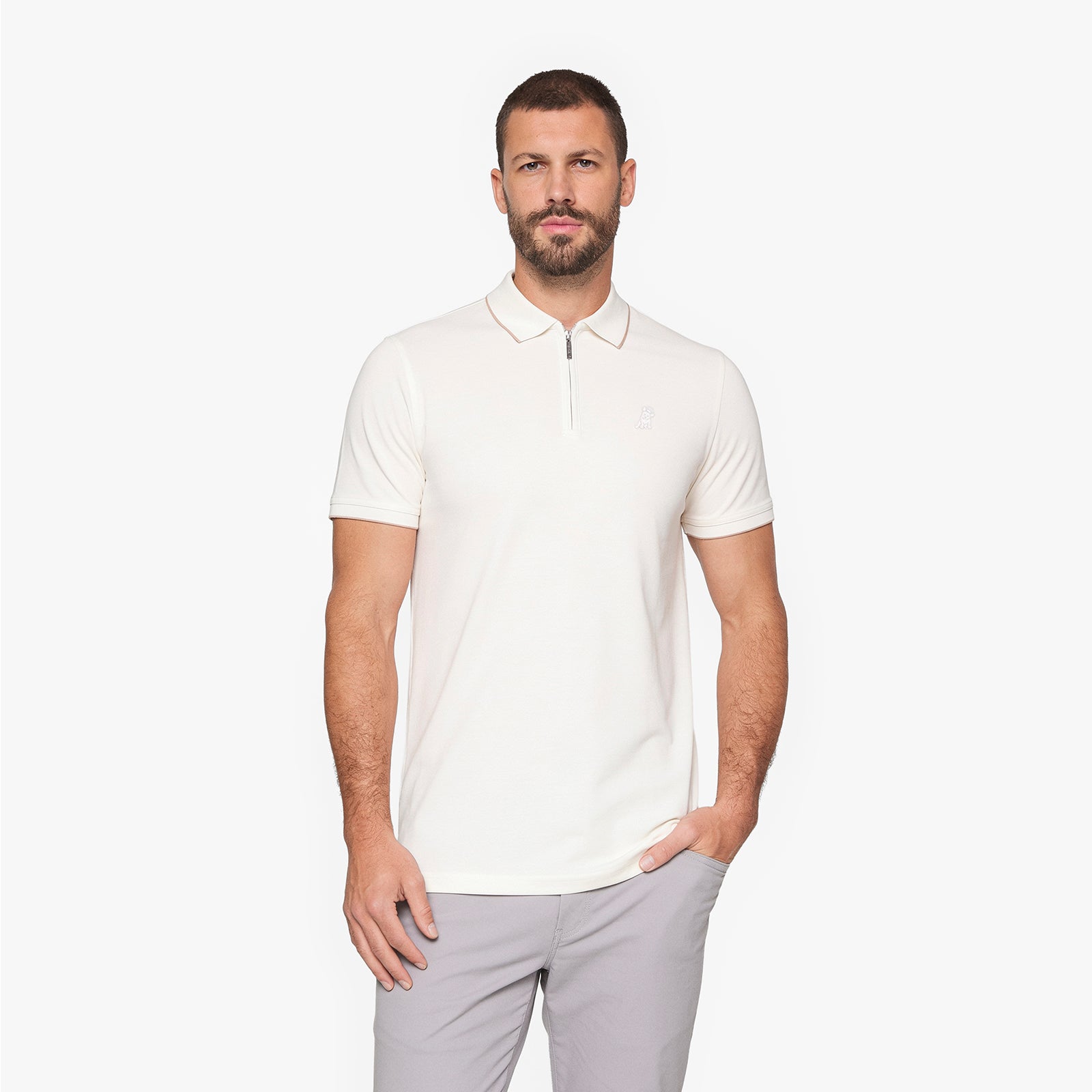 A man wearing a short-sleeve polo shirt with a half-zip collar in off-white, paired with light gray trousers, standing against a plain white studio background.