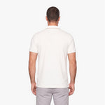 A man viewed from the back wearing a short-sleeve polo shirt in off-white and light gray trousers, set against a plain light gray studio background.