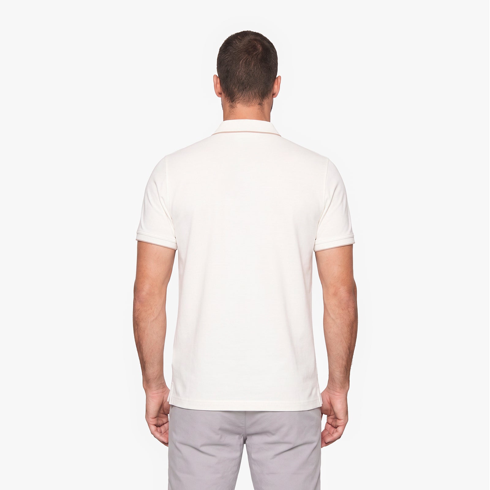 A man viewed from the back wearing a short-sleeve polo shirt in off-white and light gray trousers, set against a plain light gray studio background.