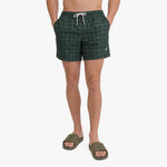 A person wearing dark green patterned swim shorts with a white drawstring and olive green slide sandals, shown from the waist down