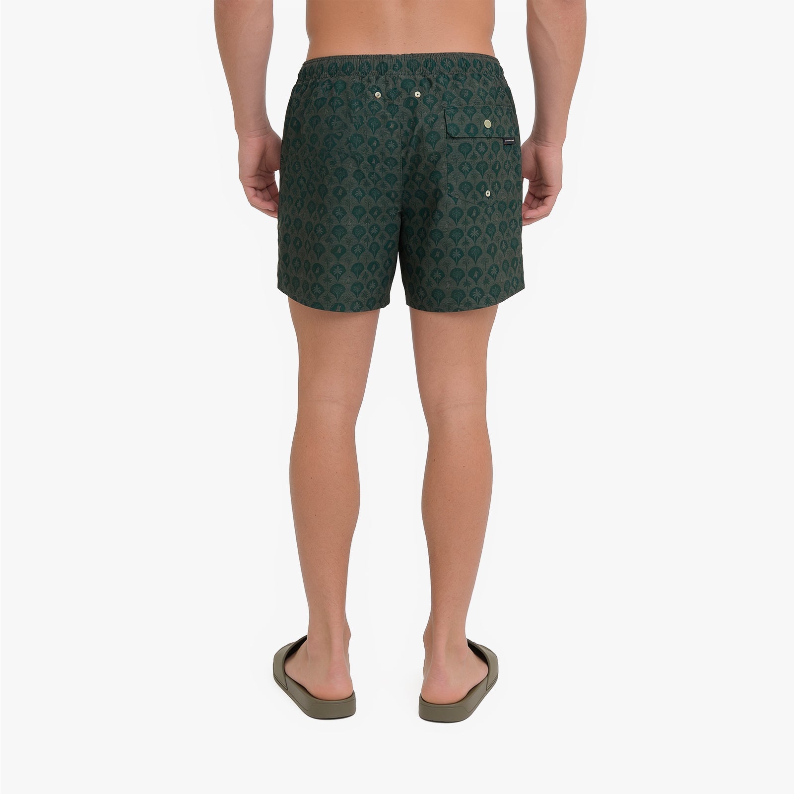 A man viewed from the back wearing dark green patterned swim trunks and olive green sandals