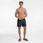 A shirtless male model stands against a light gray studio background wearing dark green patterned swim trunks with a white drawstring and olive green slide sandals.