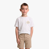 A young boy wearing a white crewneck t-shirt with a small gold and blue crest on the left chest, paired with khaki shorts, standing against a light gray background.