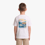 A child viewed from behind wearing a white short-sleeve T-shirt with a large multicolor graphic print on the back featuring a yacht and coastal scene, paired with khaki pants, against a light gray studio background.