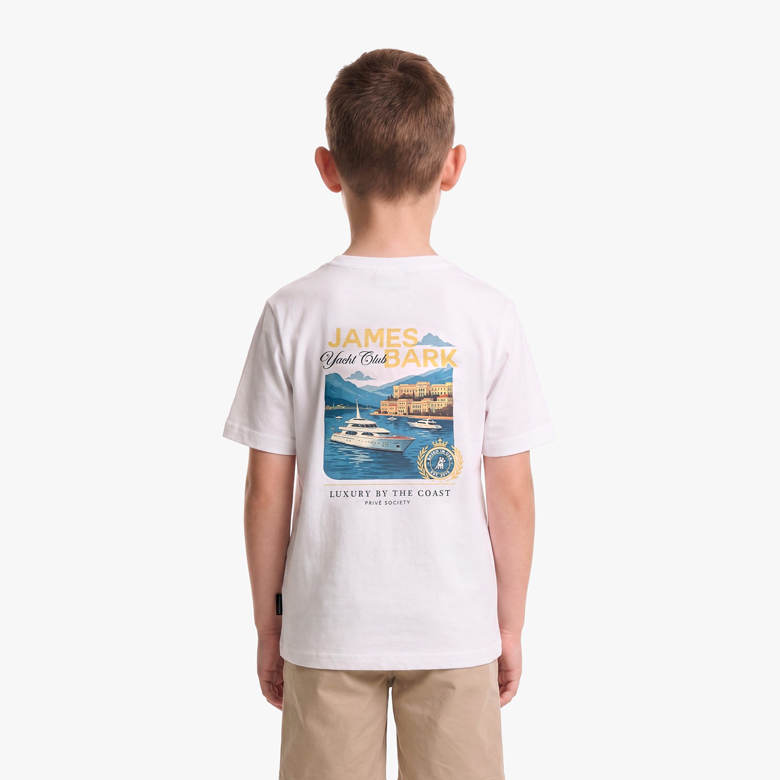 A child viewed from behind wearing a white short-sleeve T-shirt with a large multicolor graphic print on the back featuring a yacht and coastal scene, paired with khaki pants, against a light gray studio background.