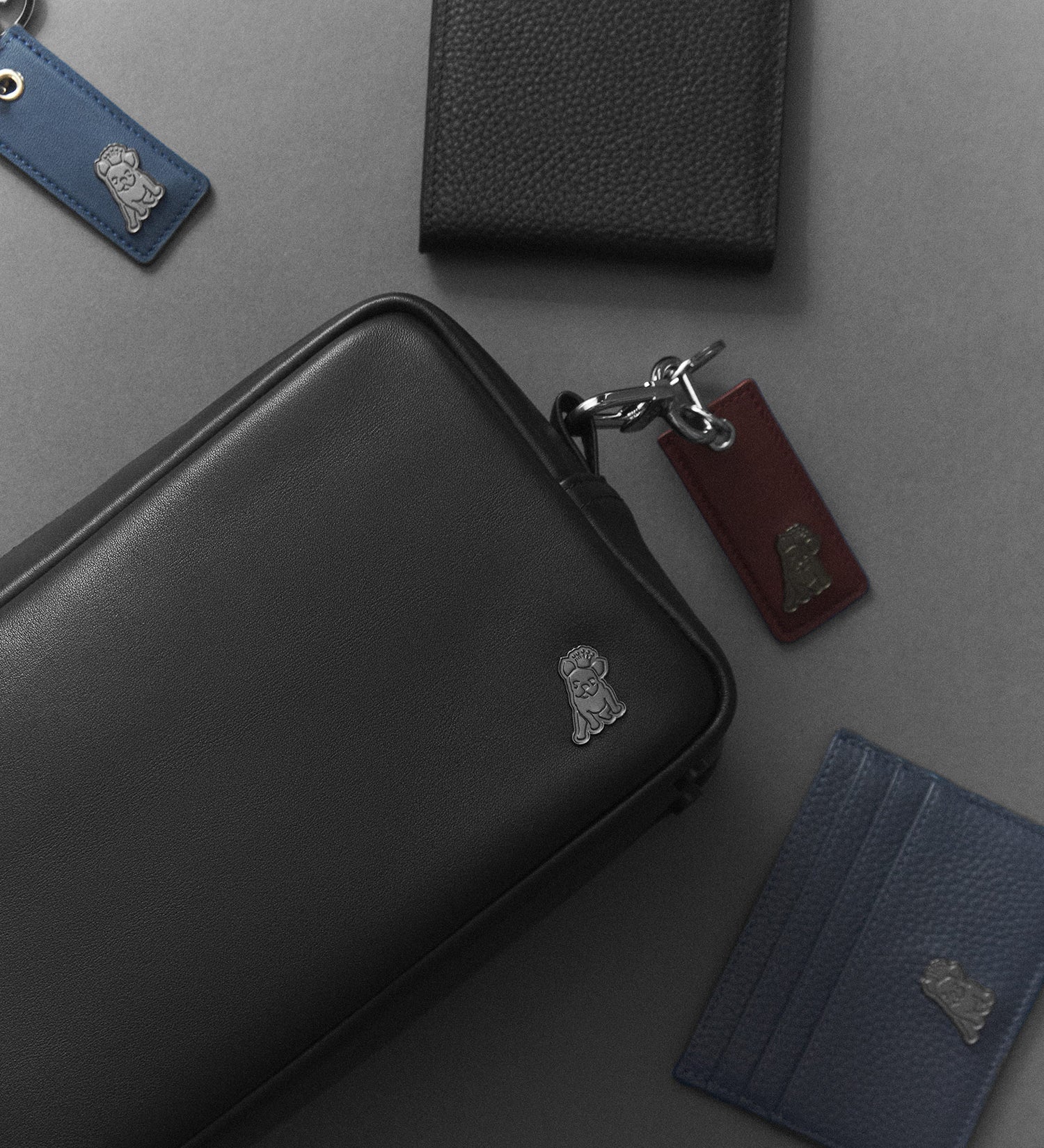 Una colección de accesorios de cuero negros y azules, incluyendo un neceser, una cartera y llaveros, dispuestos sobre una superficie gris