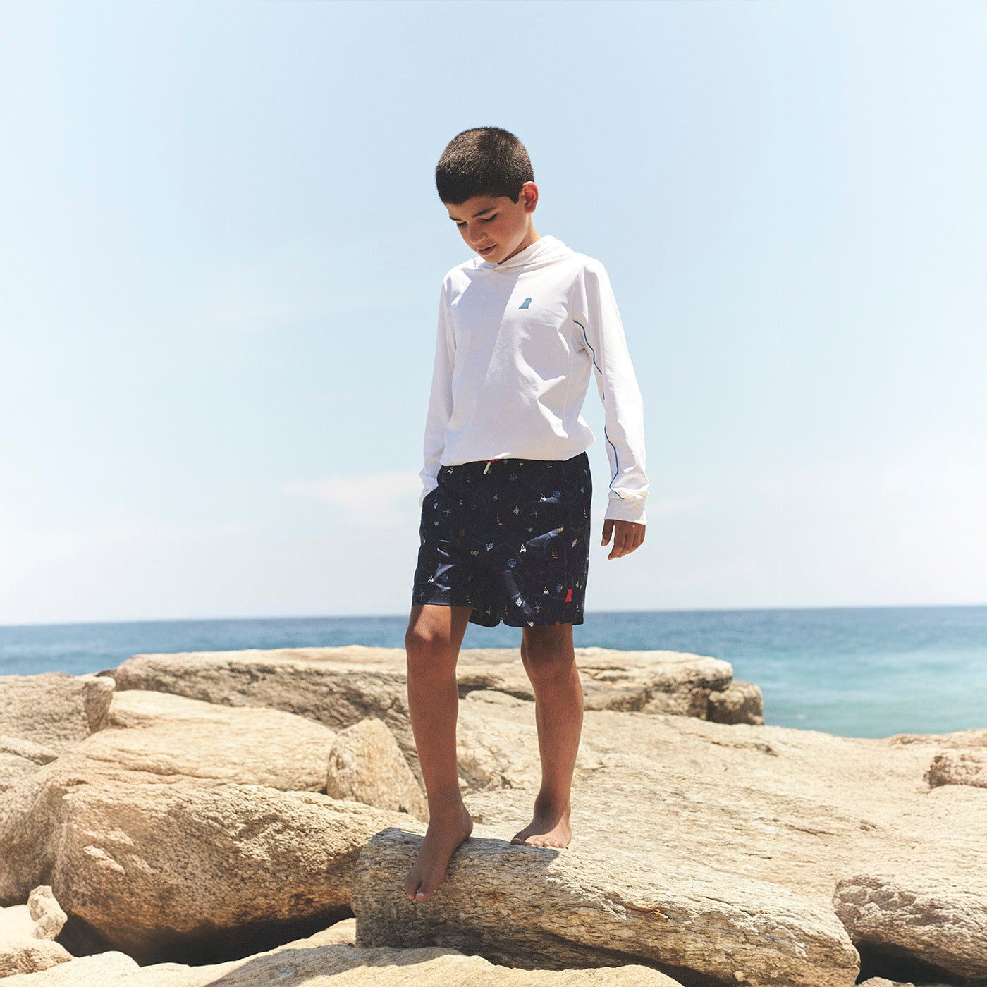 James Bark Kids Beachwear: Boys' white hooded sun shirt and navy blue patterned swim shorts in a coastal beach setting
