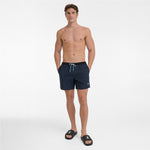 Full body view of a man wearing dark navy blue swim shorts with a black waistband and a white drawstring, paired with black sandals