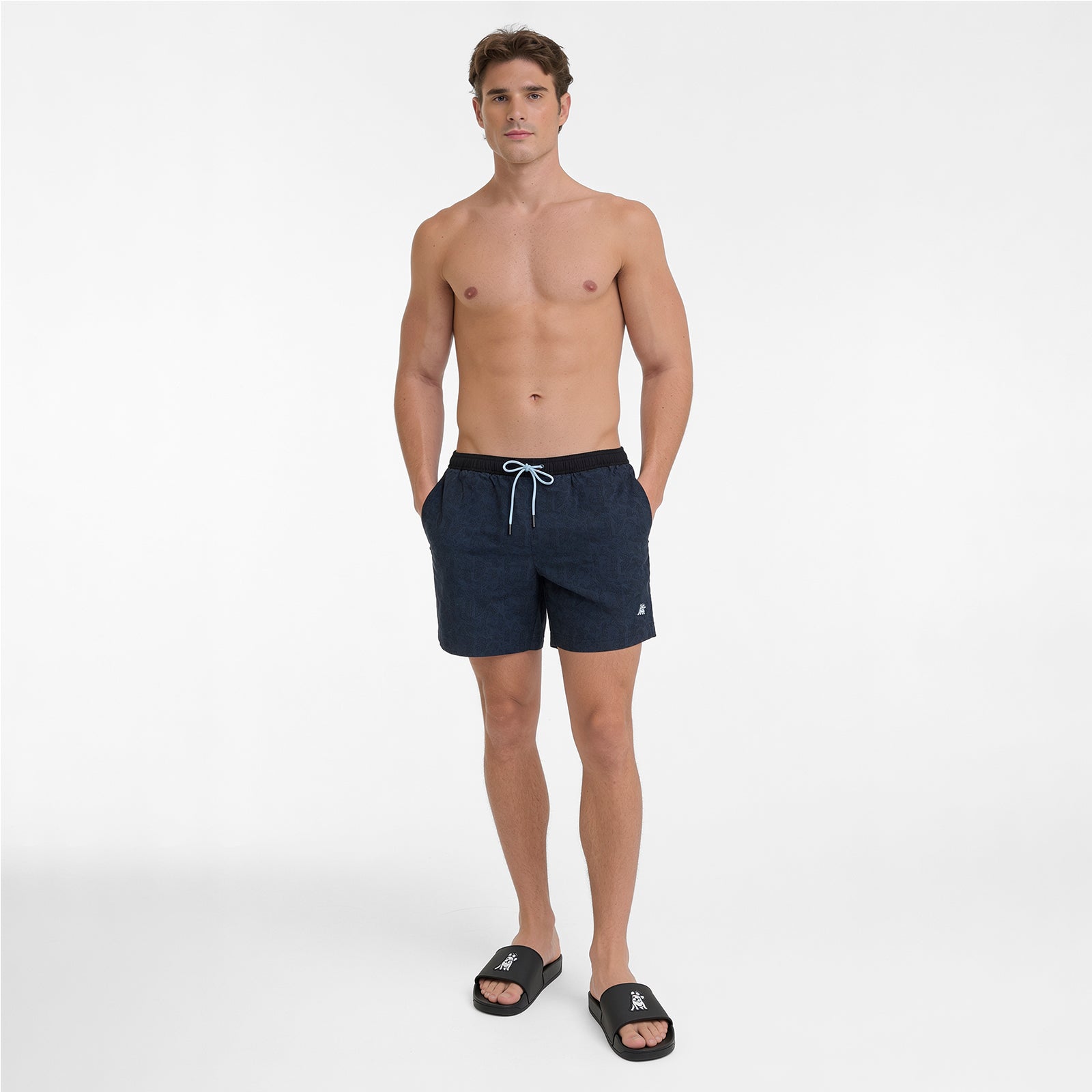 Full body view of a man wearing dark navy blue swim shorts with a black waistband and a white drawstring, paired with black sandals