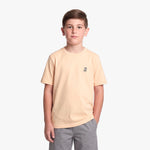 A young boy wearing a light peach T-shirt with a small logo on the left chest, paired with gray pants, standing against a white background.