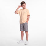 A young boy modeling a light peach T-shirt, gray shorts, and white sneakers against a white background.