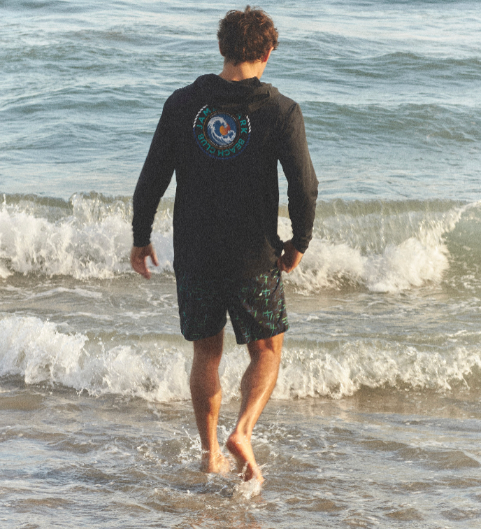 Vista trasera de un hombre con una sudadera con capucha negra y un short de baño, caminando hacia las olas del mar