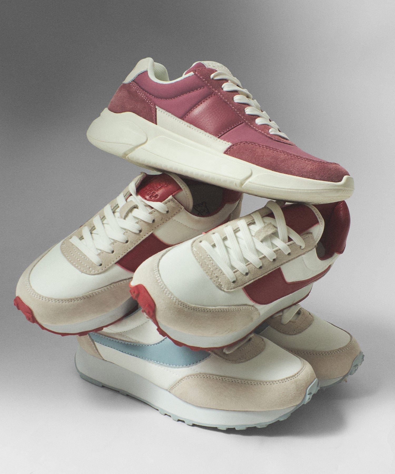  Varios pares de zapatillas de deporte en tonos rojo, azul claro y blanco, apiladas artísticamente una sobre la otra.